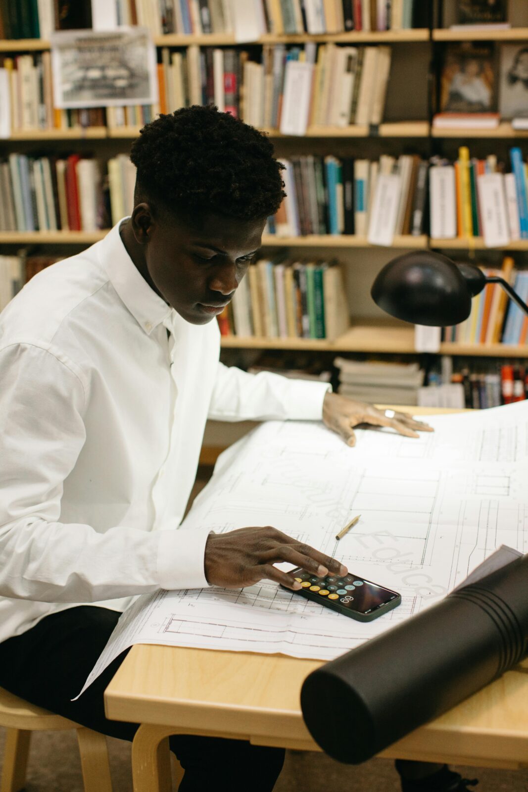 Architect in library calculating figures on large blueprints at desk.